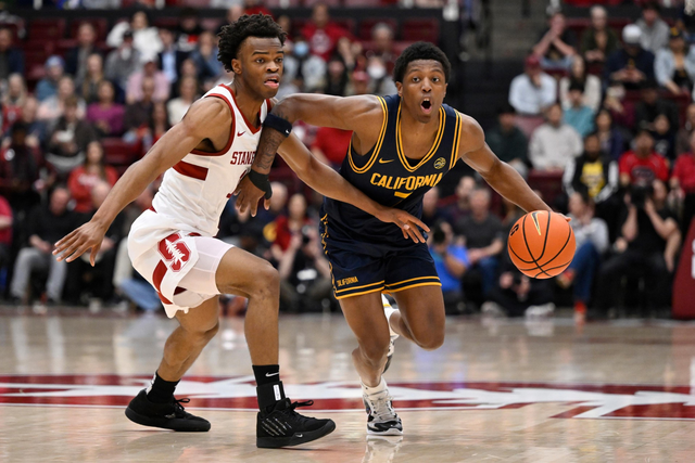 A California player dribbles past a Stanford player during a tense basketball game with spectators in the background.